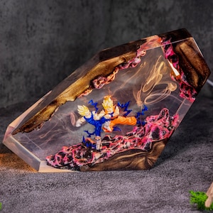May include: A clear resin lamp featuring two anime figures in a dynamic fighting pose. The base contains textured pink and black material, while the top section has a dark, metallic gold finish. Wispy, smoke-like patterns are suspended inside.