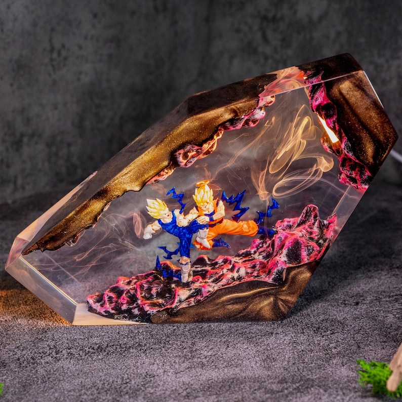 May include: A clear resin lamp featuring two anime figures in a dynamic fighting pose. The base contains textured pink and black material, while the top section has a dark, metallic gold finish. Wispy, smoke-like patterns are suspended inside.