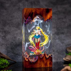 Puede incluir: Un bloque rectangular de madera pulida y resina transparente que contiene una figura de Sailor Moon. La figura tiene el pelo largo y rubio, una falda azul y un top blanco. Patrones de humo blanco están suspendidos en la resina.