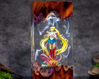 Puede incluir: Un bloque rectangular de madera pulida y resina transparente que contiene una figura de Sailor Moon. La figura tiene el pelo largo y rubio, una falda azul y un top blanco. Patrones de humo blanco están suspendidos en la resina.