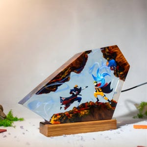 Pu&ograve; includere: Una lampada trapezoidale in legno e resina trasparente con due personaggi anime in posa di combattimento dinamica. Un personaggio indossa un mantello nero con nuvole rosse, l'altro un completo arancione e tiene un globo di energia blu.