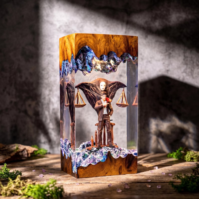 May include: A rectangular lamp made of wood and clear resin featuring a miniature figure of a man in a brown suit standing before a dark, winged mask-like structure with two hanging scales. The wood has a natural grain and textured edges.