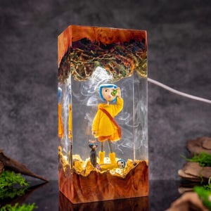May include: A rectangular resin lamp featuring a character with blue hair in a yellow raincoat and boots, accompanied by a black cat. The clear resin contains swirling white smoke effects, encased between top and bottom sections of natural wood.