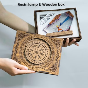 Pu&ograve; includere: Una scatola di legno con un design circolare inciso che presenta un simbolo centrale simile a una bussola circondato da lettere. All'interno, una lampada in resina mostra una miniatura in tenuta arancione che emette un raggio blu, con un cavo.