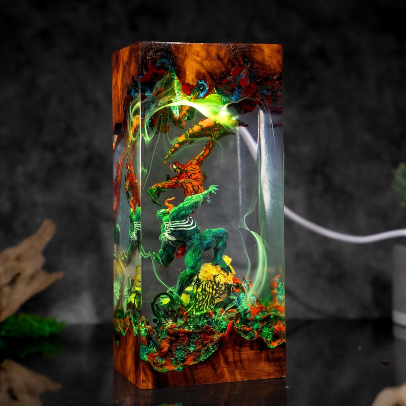 May include: A rectangular resin art piece featuring three superhero figures in vibrant colors. The figures are encased in clear resin with a wooden base and top, showcasing a dynamic scene with green, orange, and blue hues. The piece is approximately 6 inches tall.