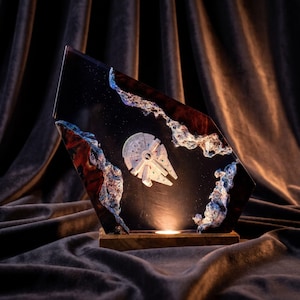 Può includere: Una lampada decorativa in resina con un'astronave Millennium Falcon bianca in miniatura incastonata in una lastra scura e traslucida. L'oggetto presenta bordi testurizzati con riflessi blu e bianchi ed è montato su una base in legno.
