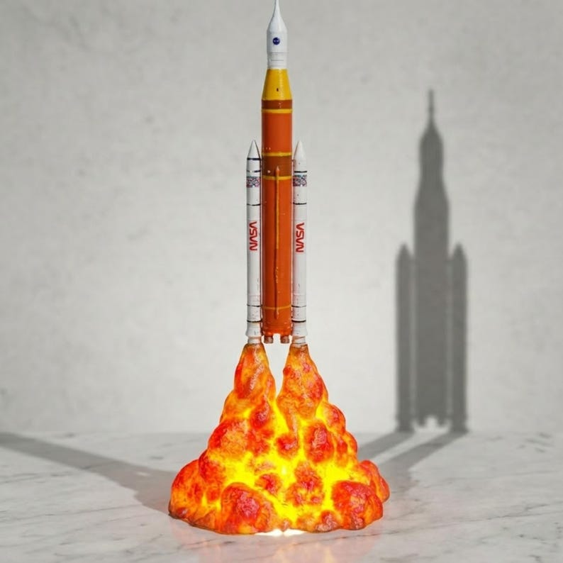 May include: A vertical model of the NASA Space Launch System rocket featuring a central orange core stage, a white upper stage, and two white side boosters. The base is illuminated with a textured, glowing orange and yellow flame effect.