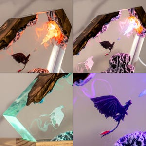 May include: A decorative resin lamp featuring a hexagonal frame with embedded dragon figures. The design includes a black dragon with red tail accents and a white dragon, set against a translucent background with internal lighting that shifts between orange, purple, and teal hues.