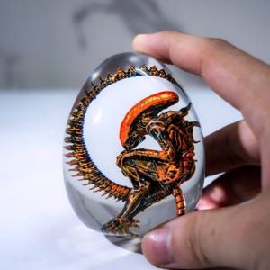 May include: A clear, egg-shaped resin sculpture featuring an orange and black alien creature. The sculpture is set against a white background within the egg, with the alien's tail wrapping around the edge. The sculpture is held by a hand.