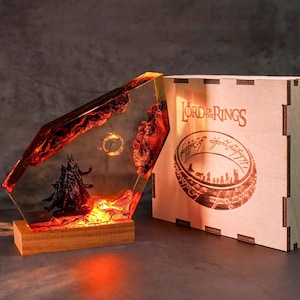 May include: A decorative lamp featuring a scene from Lord of the Rings. The lamp has a red and orange resin design with a black figure and a golden ring. It sits on a wooden base. A wooden box with the Lord of the Rings logo is next to the lamp.