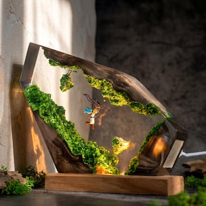 May include: A decorative lamp featuring a dark wood frame with embedded green moss and a miniature figure of a character in a white and blue outfit suspended in clear resin. The lamp sits on a rectangular wooden base and emits a warm light.