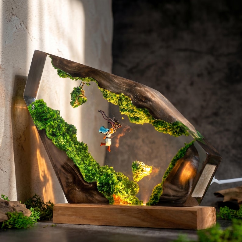 May include: A decorative lamp featuring a dark wood frame with embedded green moss and a miniature figure of a character in a white and blue outfit suspended in clear resin. The lamp sits on a rectangular wooden base and emits a warm light.