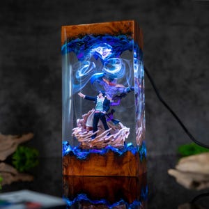 May include: A rectangular resin lamp featuring a character figurine. The lamp has a wooden base and is filled with blue and white swirling patterns. The figurine is of a person in a black coat and hat, with a purple figure behind them.