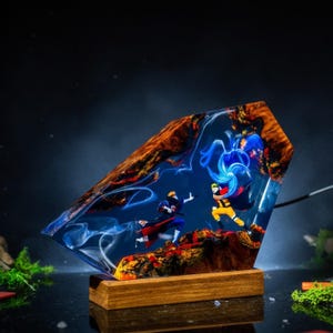 Pu&ograve; includere: Una lampada decorativa con un blocco di resina trasparente che racchiude due personaggi anime in posa da combattimento. La resina presenta effetti di luce blu vorticosi, bordi in legno naturale e una base rettangolare in legno.