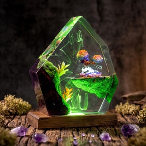 May include: A pentagonal resin sculpture featuring a translucent green interior with suspended mossy rock formations, small yellow plants, and two colorful mushroom-like structures. The piece is mounted on a rectangular wooden base.