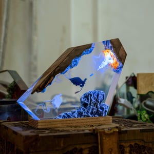 May include: A rectangular resin lamp featuring a dark wooden base and a clear resin block. Inside, a miniature dark blue dragon flies near a textured rock formation, with swirling blue smoke effects and a glowing orange and white light element.