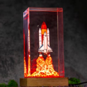 May include: A rectangular clear resin block containing a miniature space shuttle model with a white body and black accents. The base of the shuttle emits a bright orange and red glow, simulating a rocket launch, set on a light wooden base.