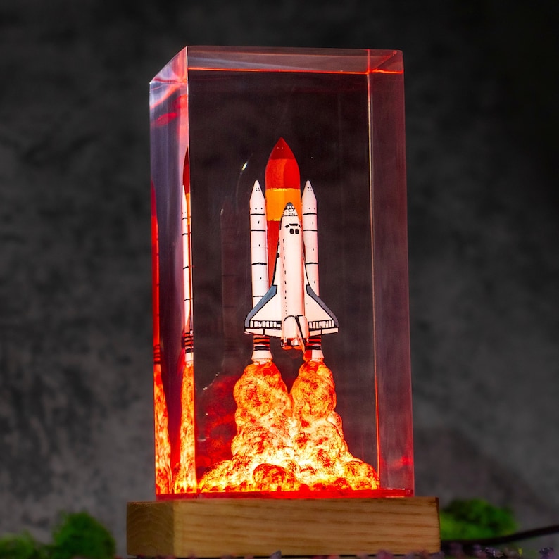 May include: A rectangular clear resin block containing a miniature space shuttle model with a white body and black accents. The base of the shuttle emits a bright orange and red glow, simulating a rocket launch, set on a light wooden base.