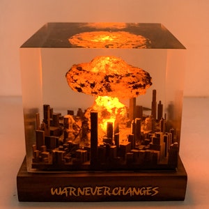 May include: A lighted sculpture depicting a nuclear explosion over a city. The explosion is orange and yellow, with a cityscape silhouette below. The base is wood, with the text "WAR NEVER CHANGES".