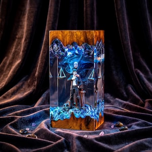 May include: A rectangular resin and wood lamp featuring a central male figure in a dark suit standing before a white mask. The interior is illuminated with blue light, showcasing swirling smoke-like patterns and hanging scales within the clear resin.