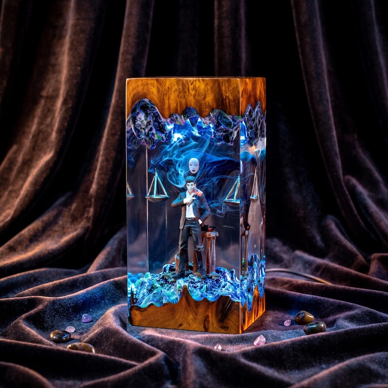 May include: A rectangular resin and wood lamp featuring a central male figure in a dark suit standing before a white mask. The interior is illuminated with blue light, showcasing swirling smoke-like patterns and hanging scales within the clear resin.