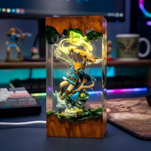 May include: A rectangular resin and wood lamp featuring a miniature figure of a blonde character in blue clothing holding a bow, suspended within clear resin. The lamp emits a warm yellow glow, illuminating the figure and a green creature base.