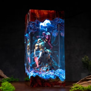 May include: A rectangular resin lamp featuring a detailed scene of a metallic armoured figure holding a glowing red sword while battling a red creature. The piece is encased in clear resin with swirling blue light effects and dark red accents.