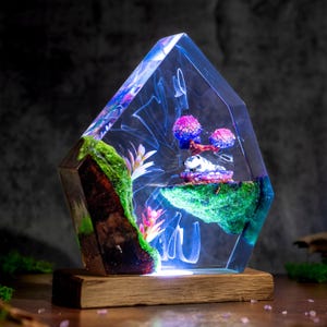 May include: A pentagonal resin sculpture featuring a miniature landscape with a wooden base, green moss, pink flowers, and a small white creature resting on a floating island. The clear blue resin contains swirling white patterns and internal lighting.