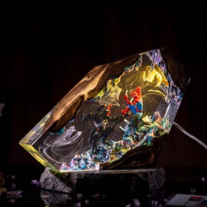 May include: A unique resin art piece featuring Spider-Man and Venom in a dynamic battle scene. The artwork is encased in a clear, geometric form with gold-colored edges, set on a dark stone base. The interior is filled with colorful, swirling details.