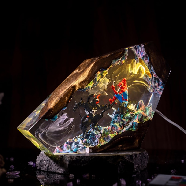 May include: A unique resin art piece featuring Spider-Man and Venom in a dynamic battle scene. The artwork is encased in a clear, geometric form with gold-colored edges, set on a dark stone base. The interior is filled with colorful, swirling details.