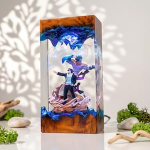 May include: A rectangular resin lamp featuring a detailed anime scene. The lamp has a wooden base and a clear resin body with blue and purple accents. The scene depicts two figures in action poses, with one figure in a black coat.