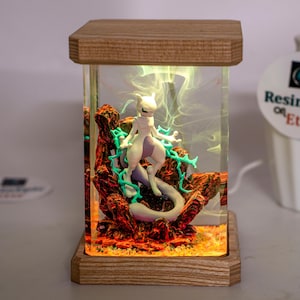 May include: A rectangular resin lamp featuring a white Mewtwo figure surrounded by teal energy effects and red rocky terrain. The clear resin block is encased between a light wood base and top, with internal lighting illuminating the scene.