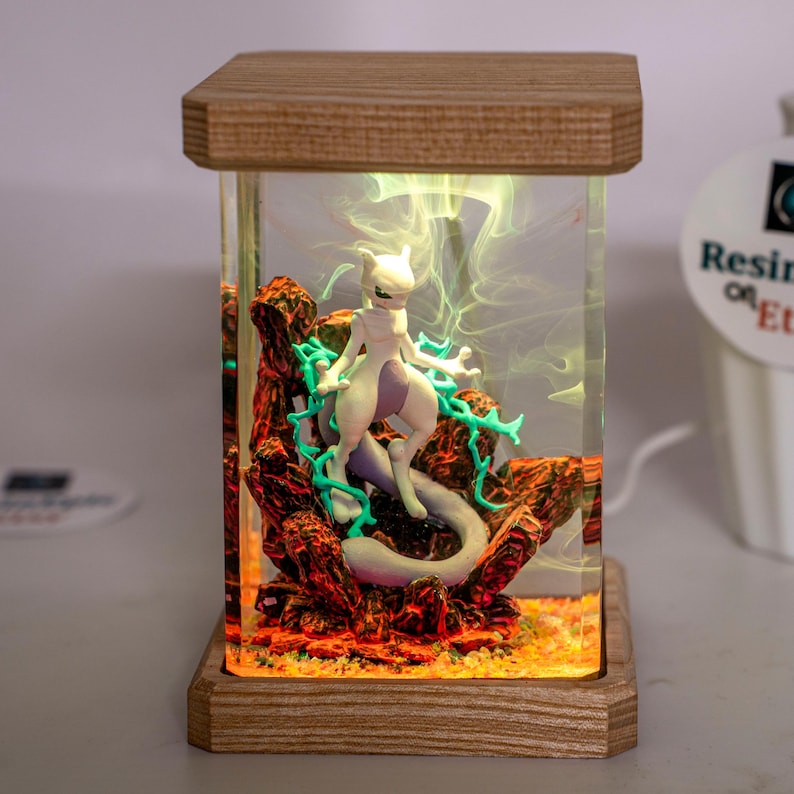 May include: A rectangular resin lamp featuring a white Mewtwo figure surrounded by teal energy effects and red rocky terrain. The clear resin block is encased between a light wood base and top, with internal lighting illuminating the scene.