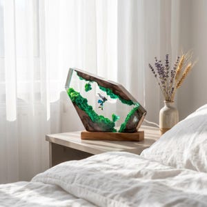 May include: A decorative lamp featuring a clear resin block with embedded dark wood, vibrant green moss, and a small figurine of a character in a blue outfit. The lamp is mounted on a rectangular wooden base and emits a soft internal light.