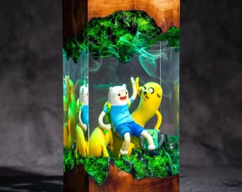 Op de afbeelding: Een rechthoekige harslamp met een basis en bovenkant van natuurlijk hout, met daarin een miniatuurscène met de Adventure Time-personages Finn en Jake. De heldere hars bevat wervelende groene rookeffecten en levendige groene mosstructuren.