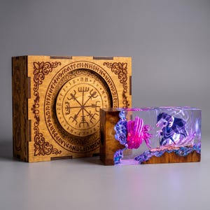May include: A square wooden box featuring an engraved circular Viking compass design and a rectangular resin block containing a miniature scene with a glowing pink creature and a dark figure, illuminated by internal purple and blue light effects.