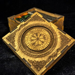 May include: A square wooden box with a removable lid featuring a laser-engraved Vegvisir symbol surrounded by a circular border of runic characters. The corners of the lid are decorated with intricate scrollwork patterns in a dark brown finish.