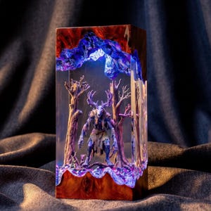 May include: A rectangular resin and wood lamp featuring a central figure with a deer skull head and antlers standing among sculpted trees. The piece is illuminated with blue and purple light, encased in clear resin with natural wood at the base.