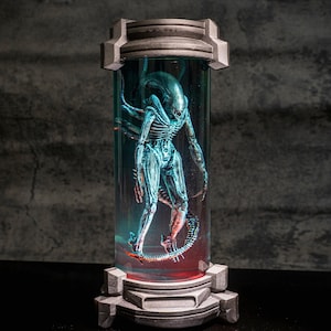 May include: A detailed sculpture of an Alien creature encased in a cylindrical glass container. The alien is a bluish-white color with a skeletal appearance. The container has a gray base and top, with a red and blue illuminated interior.