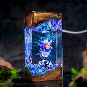 May include: A rectangular resin lamp featuring three stacked purple and black ghost-like figures suspended in a clear, swirling blue and violet liquid. The top and bottom sections of the lamp are encased in a solid, shimmering metallic gold resin.