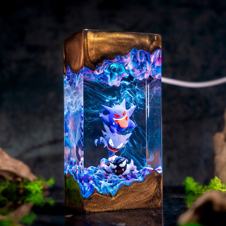 May include: A rectangular resin lamp featuring three stacked purple and black ghost-like figures suspended in a clear, swirling blue and violet liquid. The top and bottom sections of the lamp are encased in a solid, shimmering metallic gold resin.