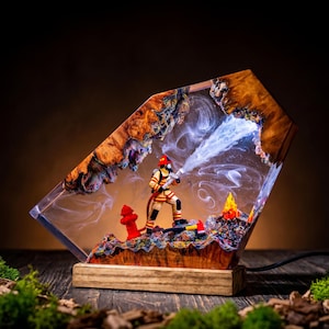 Peut inclure: Une lampe décorative représentant une figurine de pompier en tenue beige et jaune, projetant de l'eau vers un petit feu. La scène est encapsulée dans de la résine transparente avec des bords en bois naturel, sur un socle rectangulaire en bois.
