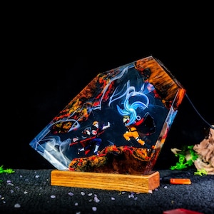 Pu&ograve; includere: Una lampada personalizzata in resina e legno con due personaggi anime in una posa di combattimento dinamica. La resina trasparente contiene effetti di fumo bianco vorticoso e accenti di legno color arancione-rosso.