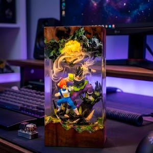 Op de afbeelding: Een rechthoekige lamp van hars en hout met twee anime-figuren in een gevechtshouding. Het heldere harsgedeelte bevat wervelende witte rookeffecten en een gloeiende gele lichtbron boven de figuren, geplaatst in een donkere houten basis.