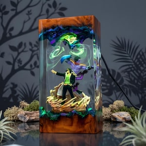May include: A rectangular resin lamp featuring a detailed diorama of anime characters. The lamp has a wooden base and top, with a clear resin body. The figures are set on a coral-like base, with green and purple accents. The lamp is lit with green and blue lights.