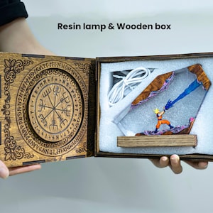 Op de afbeelding: Een houten doos met een harslamp en een figuurtje in een oranje en blauw pak dat een blauwe energiestraal afvuurt. Het deksel heeft een ingewikkeld cirkelvormig ontwerp met runen. Tekst: Resin lamp & Wooden box.