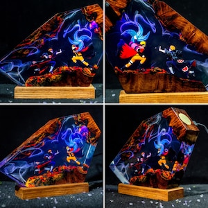 Pu&ograve; includere: Una lampada decorativa con base in legno e un blocco di resina contenente due figure anime in posa da combattimento. La resina presenta effetti di fumo blu vorticoso e uno sfondo scuro, con le figure che indossano abiti neri e arancioni.