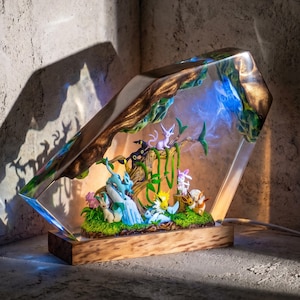 Peut inclure: Une lampe en résine trapézoïdale présentant une collection de figurines miniatures d'évolutions d'Évoli nichées dans une scène forestière. La résine transparente renferme une branche d'arbre, du feuillage vert et de petites créatures.