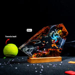 Pu&ograve; includere: Una lampada decorativa in resina e legno con una dinamica scena di battaglia tra due personaggi anime, uno con mantello nero e l'altro in tenuta arancione con un vortice di energia blu. L'oggetto &egrave; etichettato Size L accanto a una pallina da tennis.