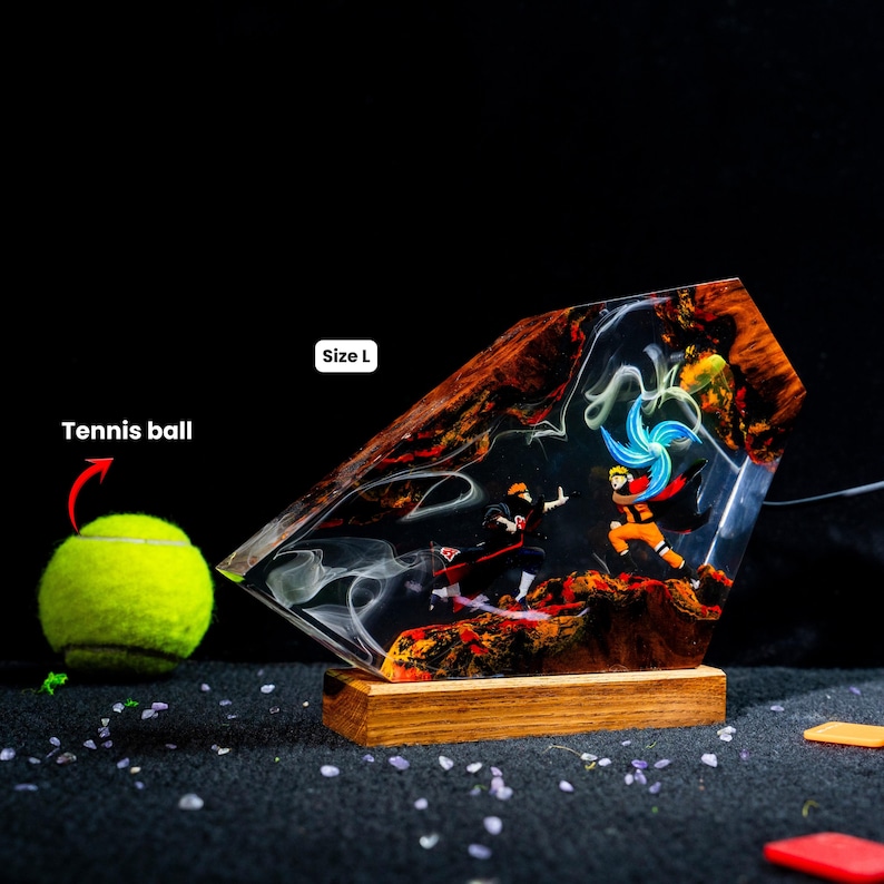Pu&ograve; includere: Una lampada decorativa in resina e legno con una dinamica scena di battaglia tra due personaggi anime, uno con mantello nero e l'altro in tenuta arancione con un vortice di energia blu. L'oggetto &egrave; etichettato Size L accanto a una pallina da tennis.